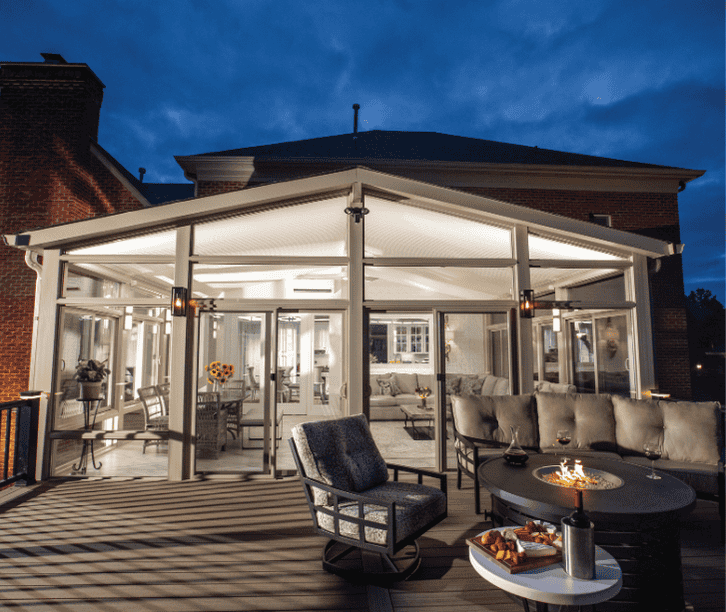 Modern four season sunroom addition with expansive windows, connected deck seating area, and outdoor fire table at dusk.