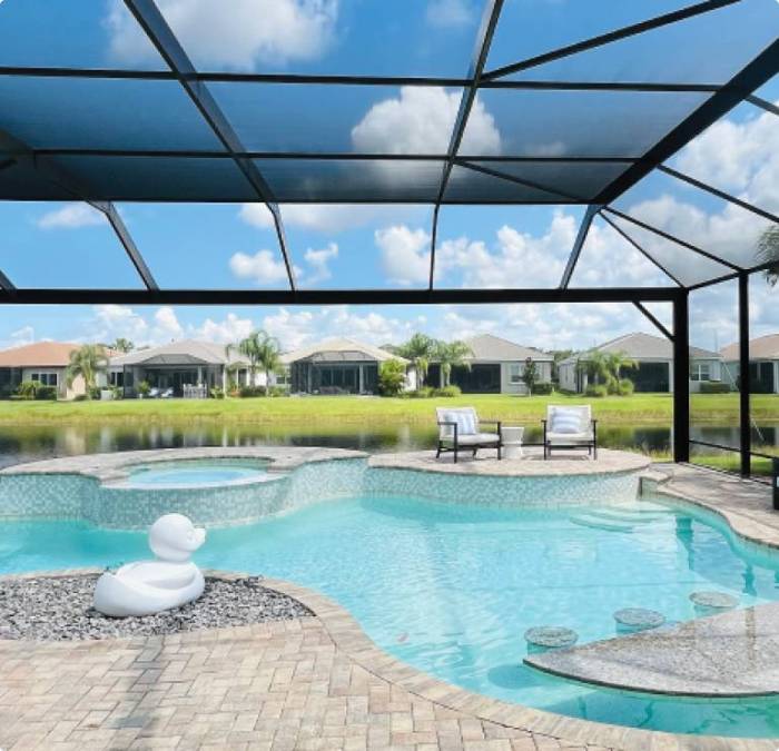Screen-enclosed backyard pool and spa overlooking a scenic pond, featuring a modern patio with lounge chairs and playful duck float—designed for stylish relaxation and year-round outdoor enjoyment.