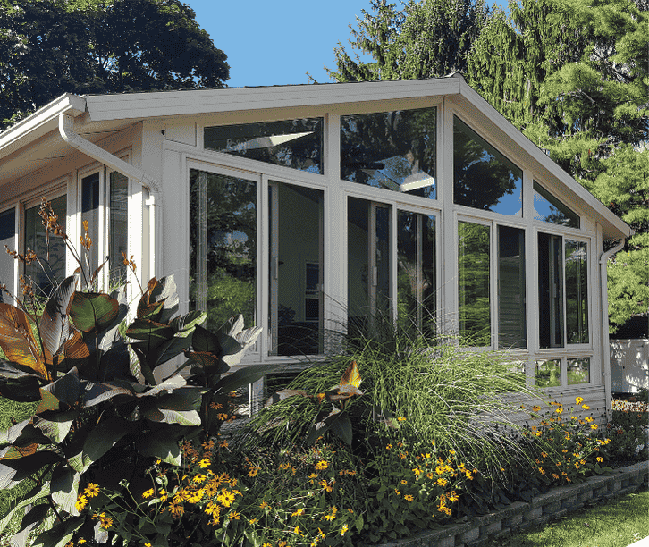 White four season sunroom with large glass windows and sloped roof surrounded by colorful garden landscaping.