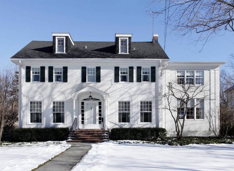 Classic two-story white colonial-style home with black shutters and a symmetrical facade, set against a clear winter sky with snow-covered lawn—showcasing timeless curb appeal and elegant design.