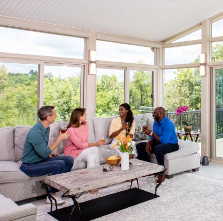 Four people relaxing and enjoying conversation in a bright, modern sunroom with large floor-to-ceiling windows and scenic views—showcasing a completed home addition that blends comfort with style.