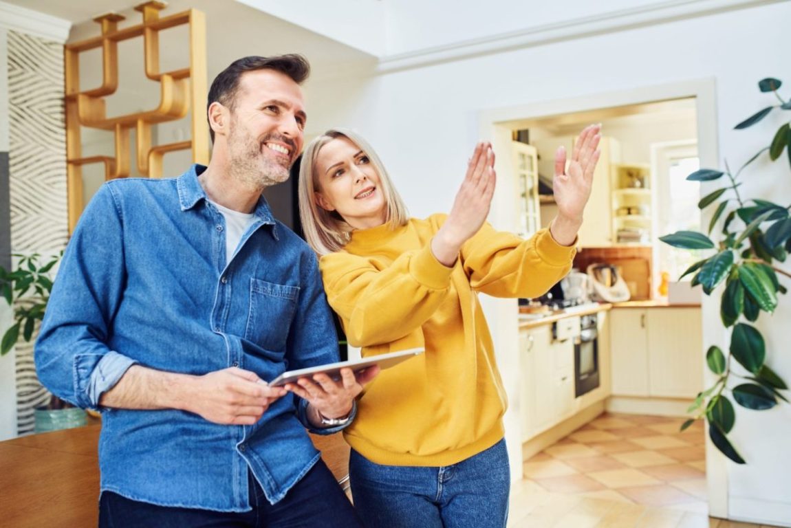 A middle-aged couple holding a tablet stand in the middle of their house smiling, gesturing, and appearing to discuss a home improvement project.