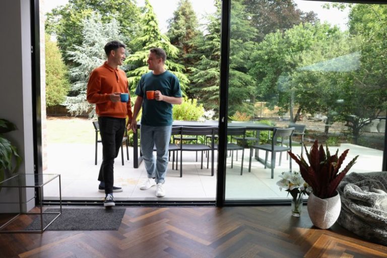 Two men are on the outdoor patio of their home, holding mugs as they walk indoors.