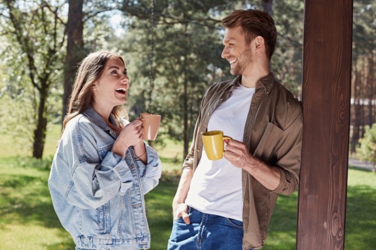A happy couple stands on their porch laughing and talking with coffee in hand.