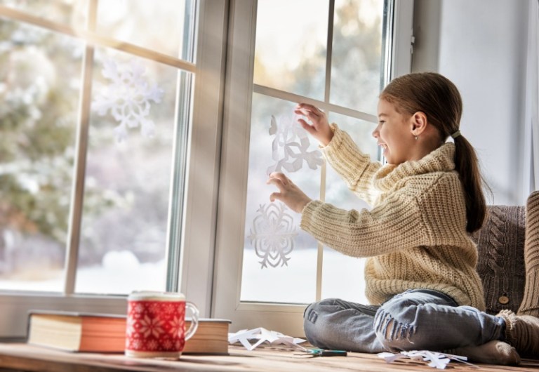 A young girl smiles as she hangs paper snowflakes in the window overlooking a snowy yard.