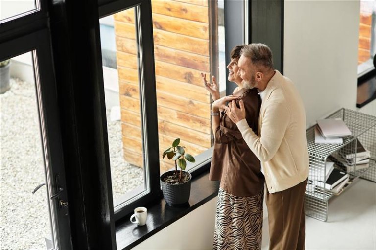 An older couple smiles and talks to each other while embracing and looking out of the window of their home.