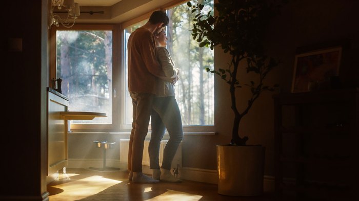 A happy couple embraces as they stand in a sunbeam while looking out the window from their kitchen.