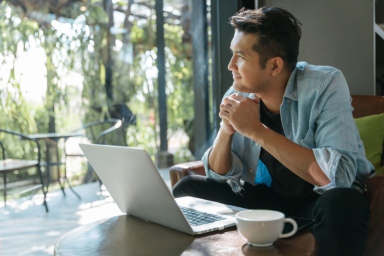 A man sits in front of his laptop in his living room while looking outside at his backyard.