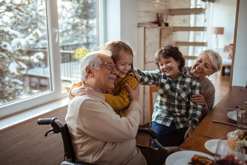 An elderly grandfather in a wheelchair and grandmother hug their grandkids at their dining table.