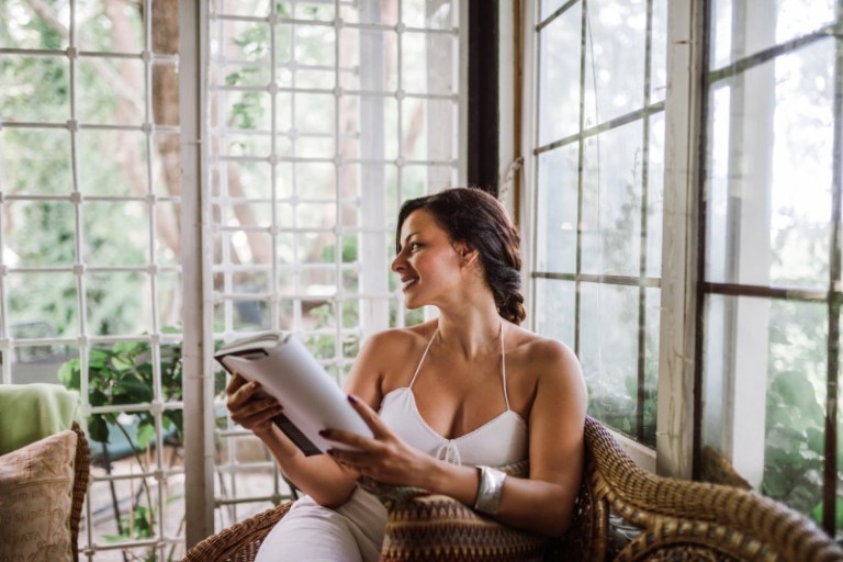 A woman in a summer dress reads a book in a sunroom filled with natural light.