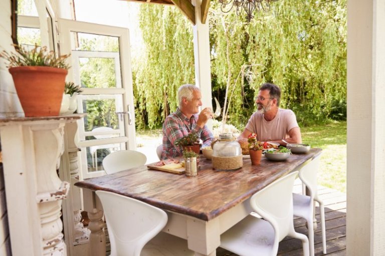 Two older men enjoy a meal at a table on a patio at a beautiful home.