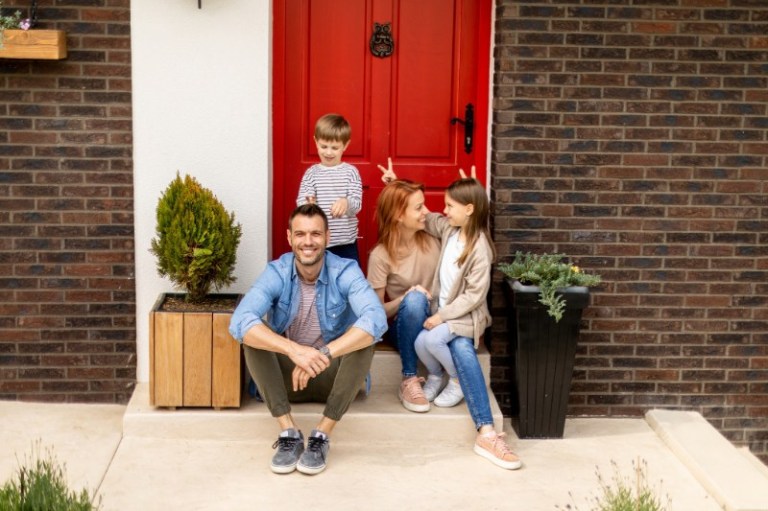 Fun and silly family sit in front of the bright red front door of their home.