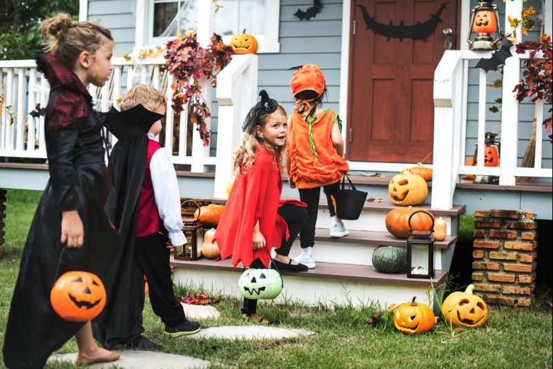 Three young kids and one older girl are dressed in Halloween costumes to trick-or-treat at a neighbor's front door with pumpkin candy pails.