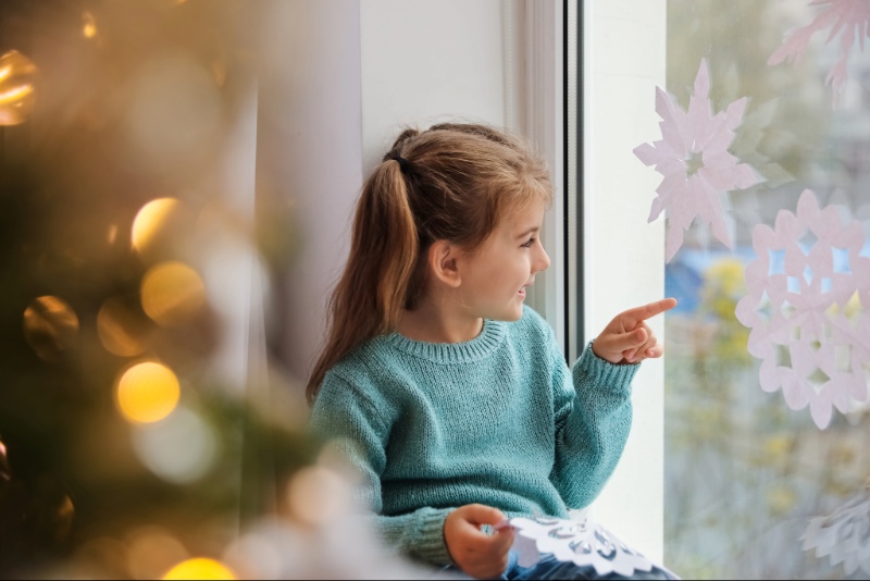A young girl sits in a big window pointing at paper snowflake cutouts clinging to the glass.