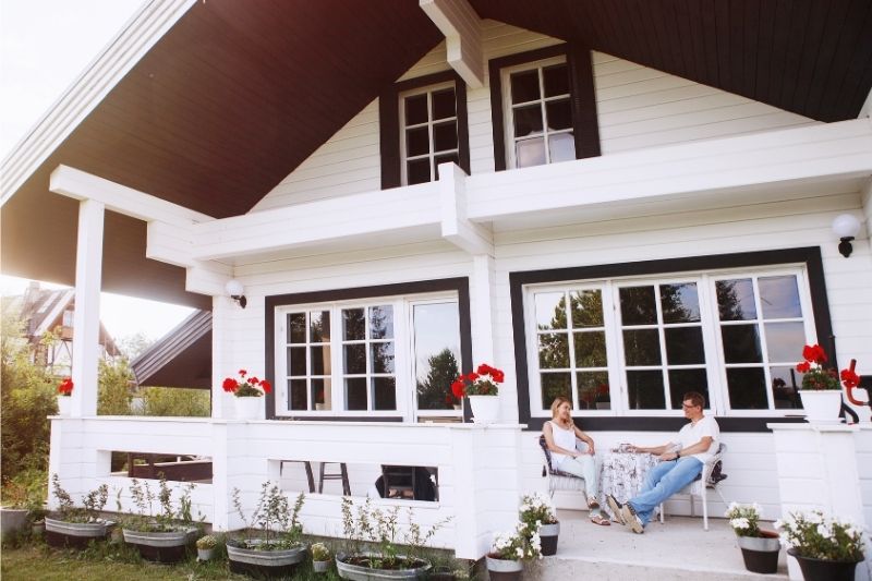 Couple sitting on front porch of a renovated house with large gable roof, white siding, and black trim—highlighting architectural style considerations for home renovations.