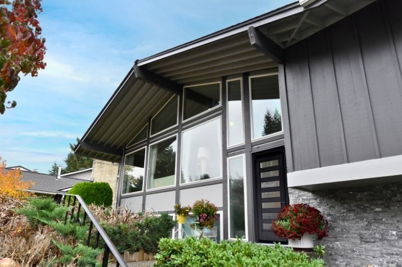 Mid-Century Modern house with low-pitched roof, large floor-to-ceiling windows, gray siding, and stone exterior accents.