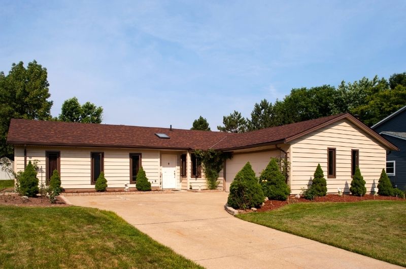 Single-story ranch style house with beige siding, brown roof, attached garage, and simple, but elegant front yard landscaping.