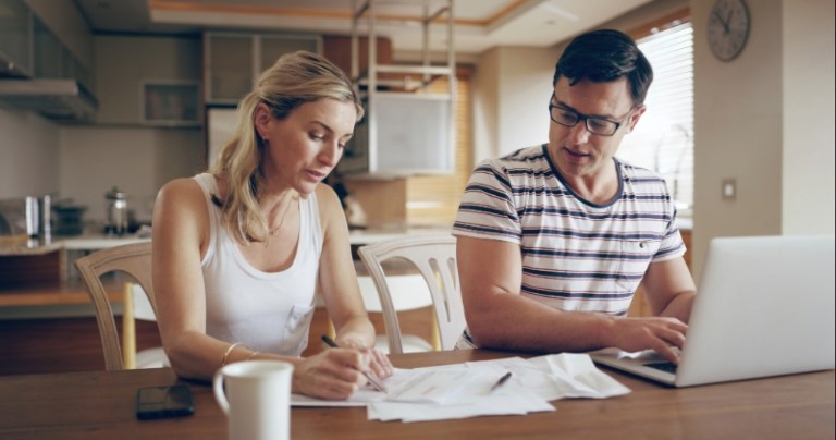 A couple sits at the table in their dining room with a laptop and papers having a calm and thoughtful discussion.