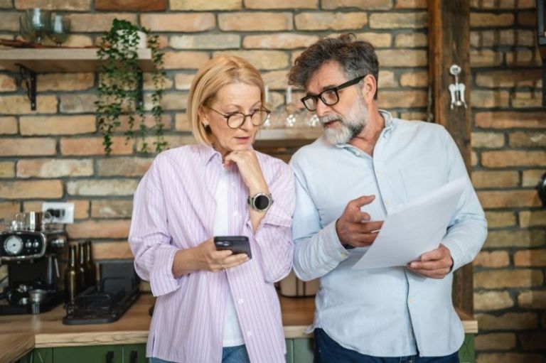 A middle-aged couple stand in their kitchen looking at papers and a phone having a discussion.