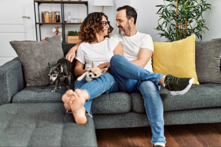A happy middle-aged couple sits together on a couch with their two small dogs.
