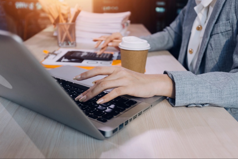 A woman uses a laptop and calculator at a desk with a coffee cup at her side.