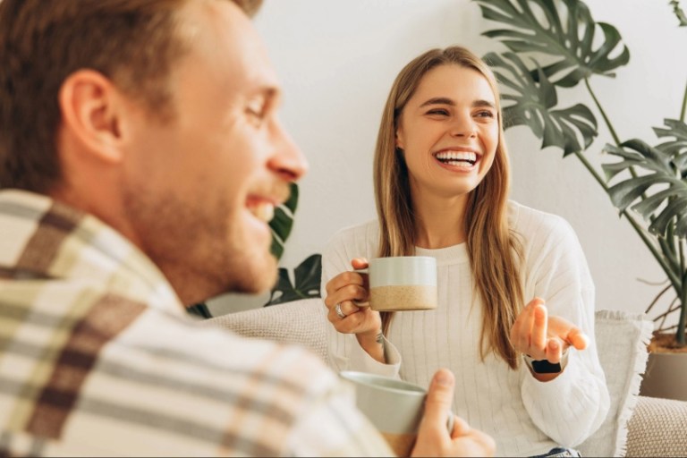 A man and woman laugh while having coffee on a couch in a living room.