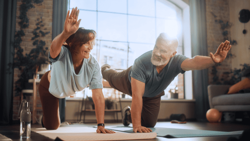 A happy middle age couple does yoga together in their home.