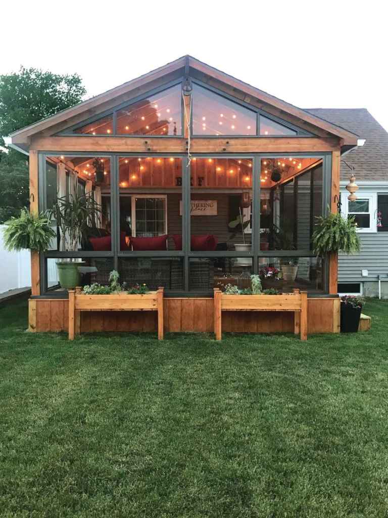 Looking in from the outside of a full glass wall sunroom with raised bed plants and string lights.